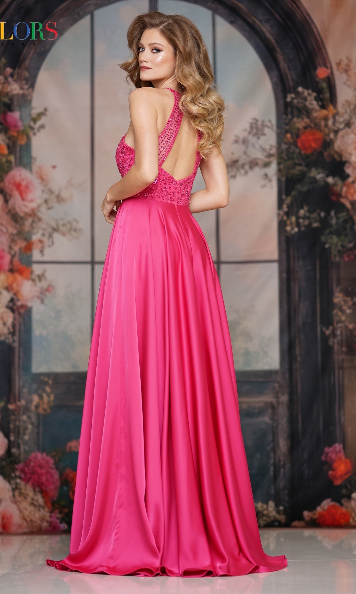 A woman with long, wavy blonde hair wears the Colors Dress 3822 high-neck halter prom gown by Colors Dress in vibrant pink with a beaded bodice, standing before an arched window and floral backdrop.